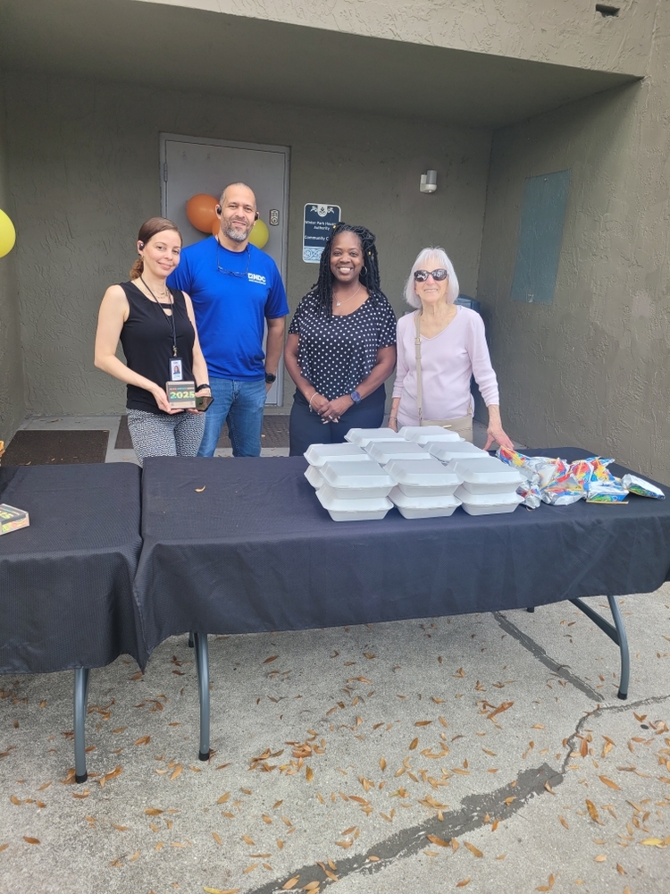 Four people standing behind a table with to go lunch containers.