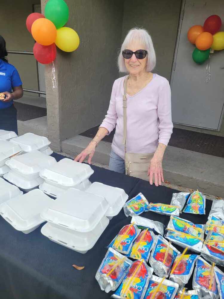 A lady standing behind a table holding containers and drinks the Luncheon.